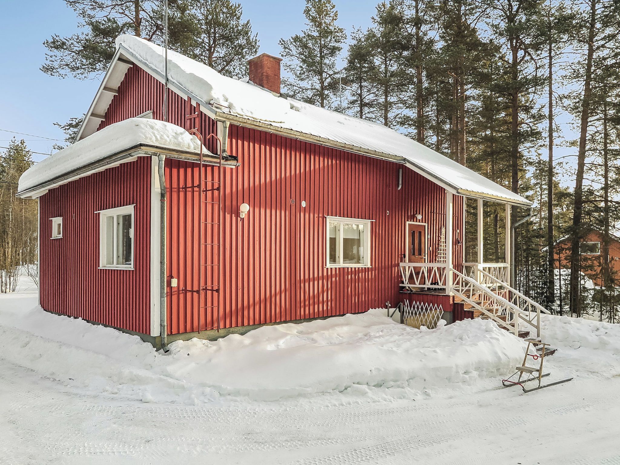 A red wooden cabin with a snow-covered roof, a front porch, and a staircase leading to the entrance.
