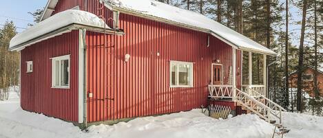 A red wooden cabin with a snow-covered roof, a front porch, and a staircase leading to the entrance.