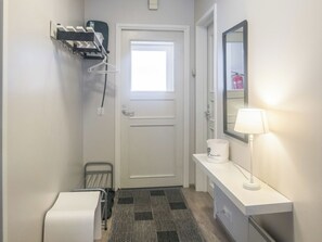Habitación de hotel con una puerta blanca, un ventanal pequeño, una mesita de noche blanca con una lámpara, una silla blanca y un perchero metálico con ganchos.