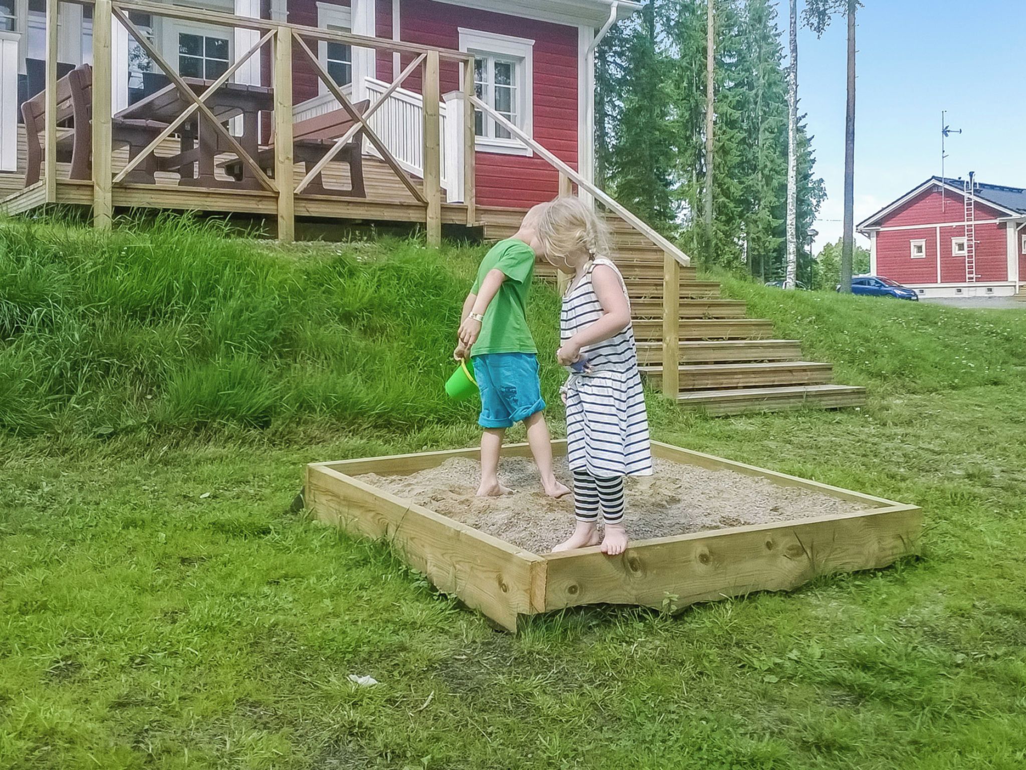 Zwei Kinder spielen in einem erhöhten Blumenbeet mit einer Holzterrasse, im Hintergrund ein rotes Gebäude.