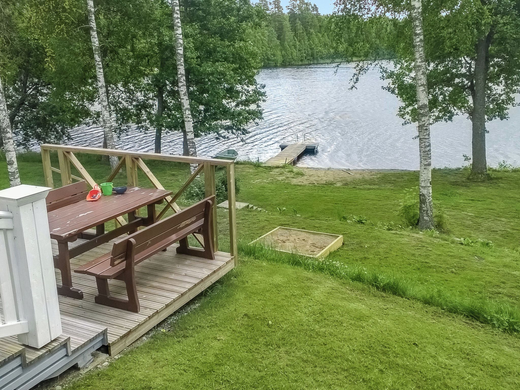 Puinen terassi, jossa on piknikpöytiä, ja pieni vene, joka on ankkuroitu tyynen, puiden ympäröimän järven rantaan.