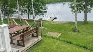A wooden deck with picnic tables and a small boat docked by a calm lake surrounded by trees.