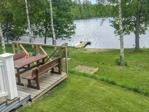 Une terrasse en bois avec des tables de pique-nique et un petit bateau amarré au bord d’un lac paisible, entouré d’arbres.