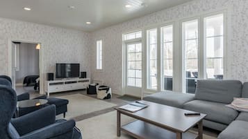 Ein geräumiges Wohnzimmer mit einem Flachbildfernseher, grauen Sofas, einem Couchtisch und großen Fenstern.