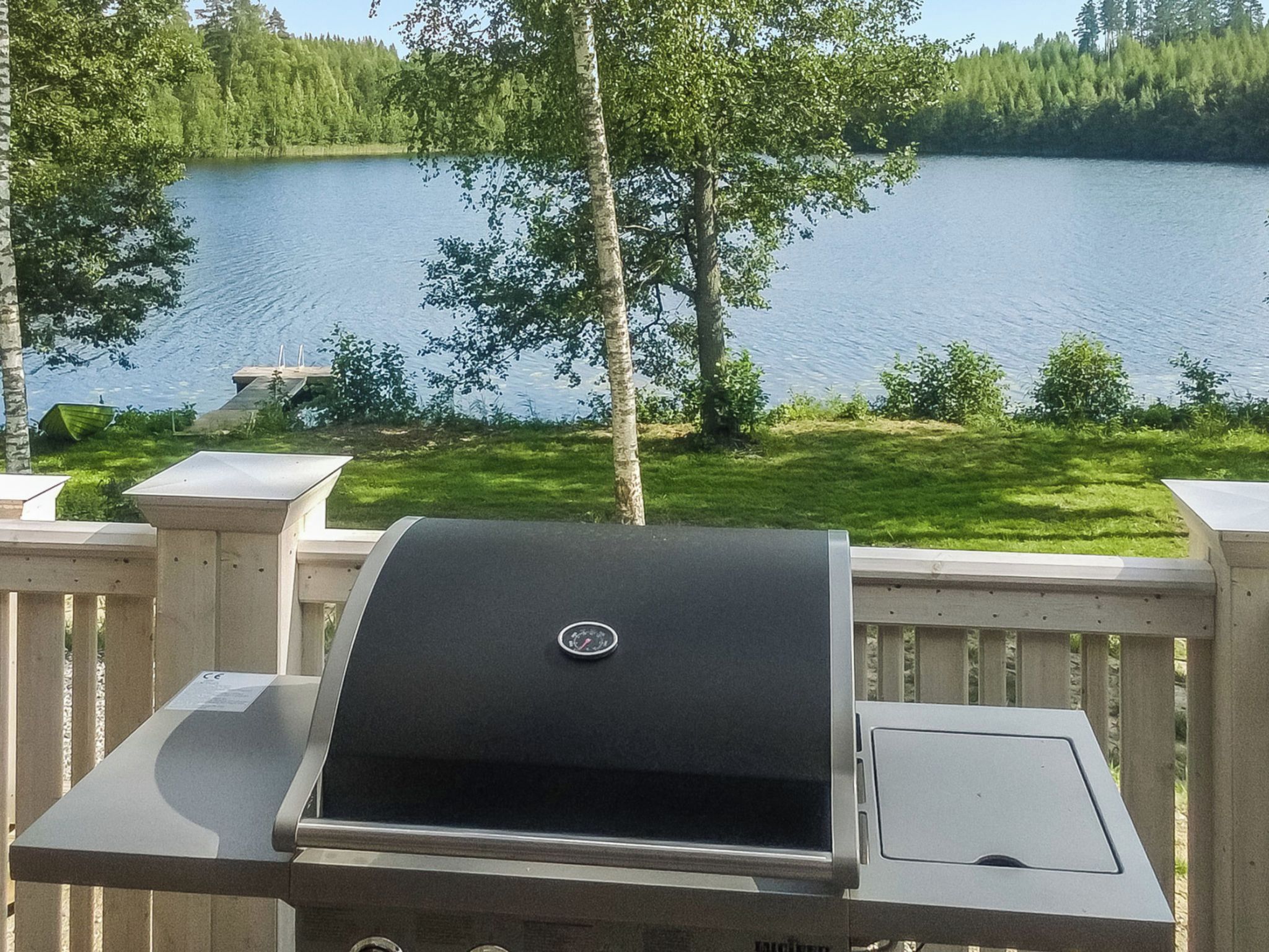 A grill on a deck overlooking a lake with trees and a dock in the background.