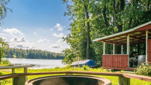 Una cabaña junto al lago con un hogar a la leña de hormigón, una terraza de madera y una zona de estar cubierta con vistas al agua.