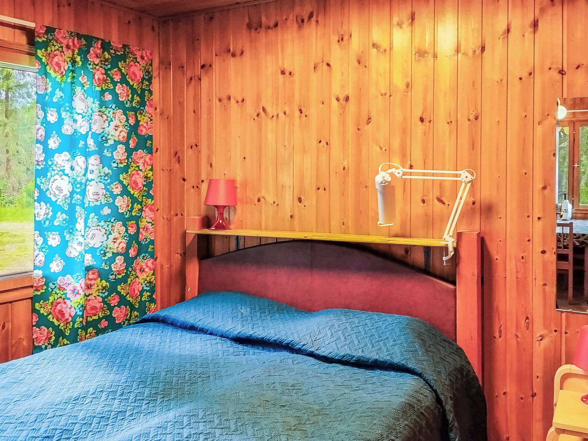 Ett rum med träpanel, en säng, ett nattduksbord, en lampa och ett fönster med blommiga gardiner.