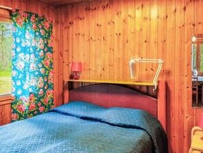 Ein Zimmer mit Holzboden, einem Bett, einem Nachttisch, einer Lampe und einem Fenster mit blumigen Vorhängen.