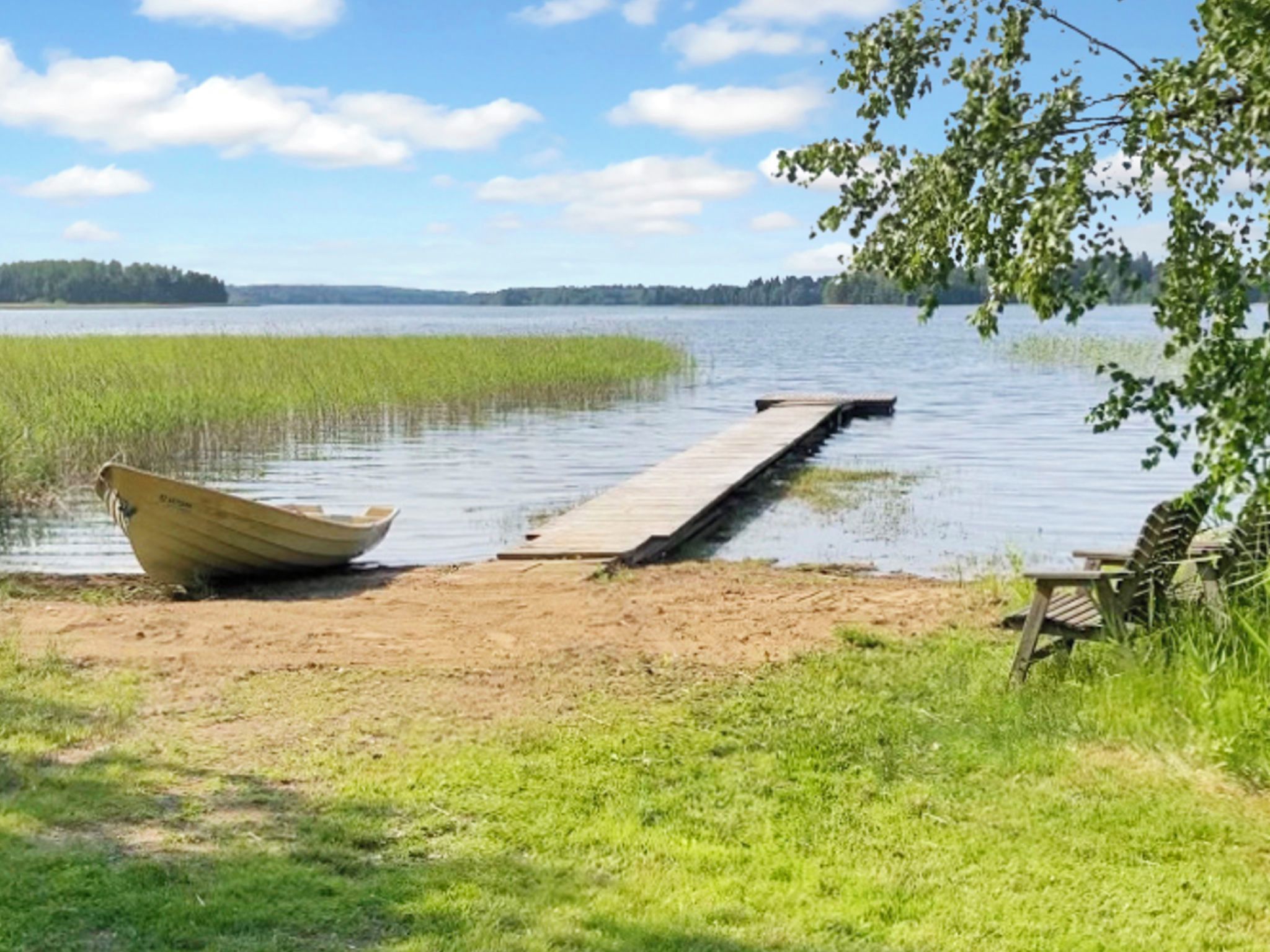 A calm lake with a wooden dock extending into the water, a boat on the shore, and a bench under a tree.