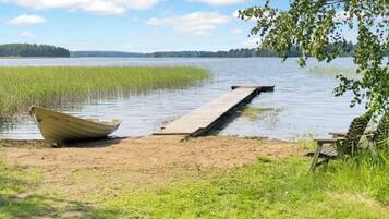 A calm lake with a wooden dock extending into the water, a boat on the shore, and a bench under a tree.