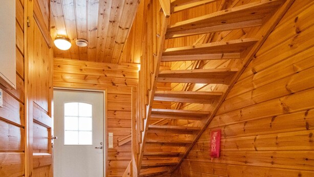 Interior de madera con una escalera que lleva a un nivel superior, una puerta con ventanal y un extintor en la pared.