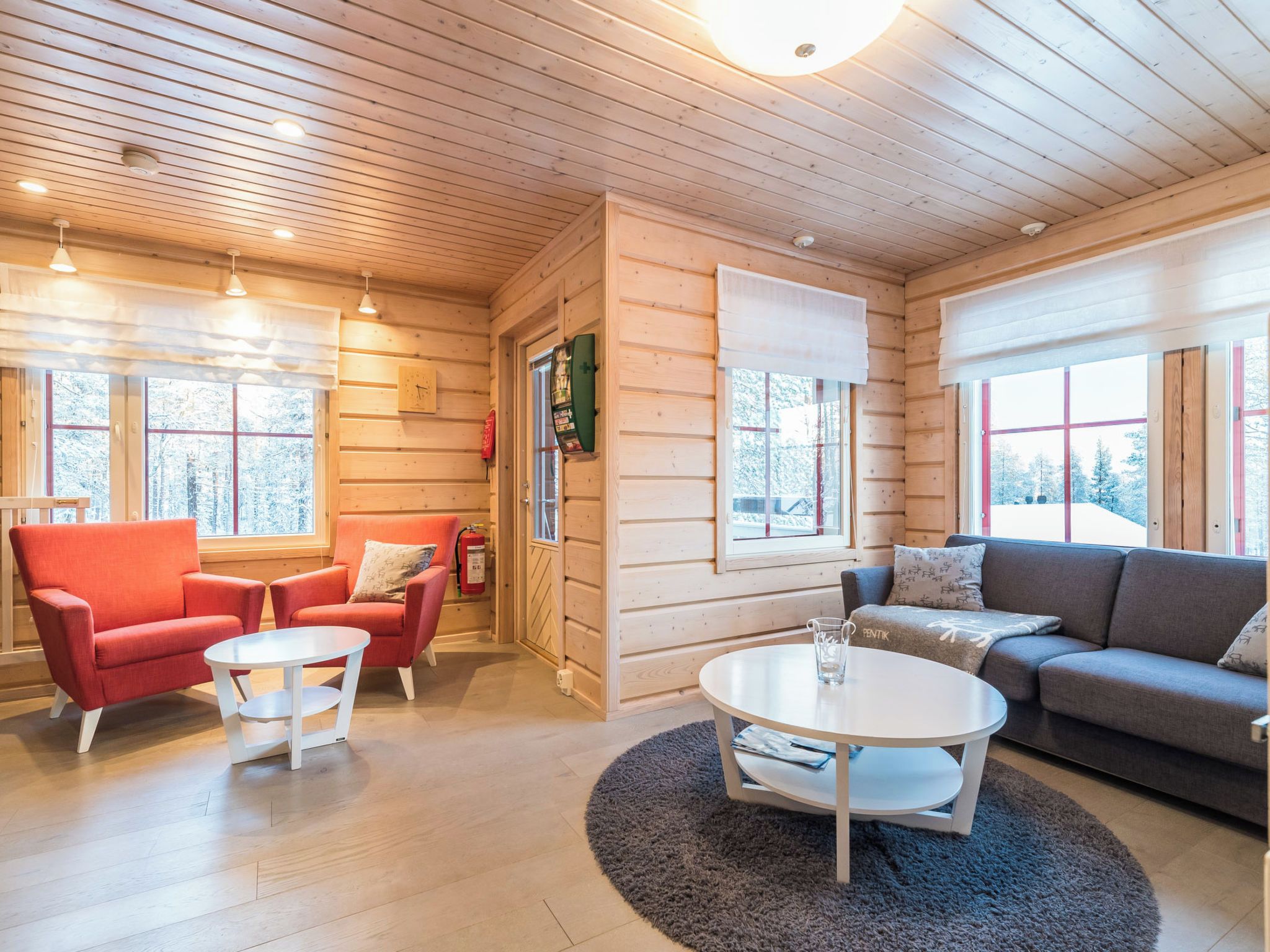 A log cabin interior with a living area, two red armchairs, a grey sofa, a round coffee table, and large windows with a view of snow-covered trees.