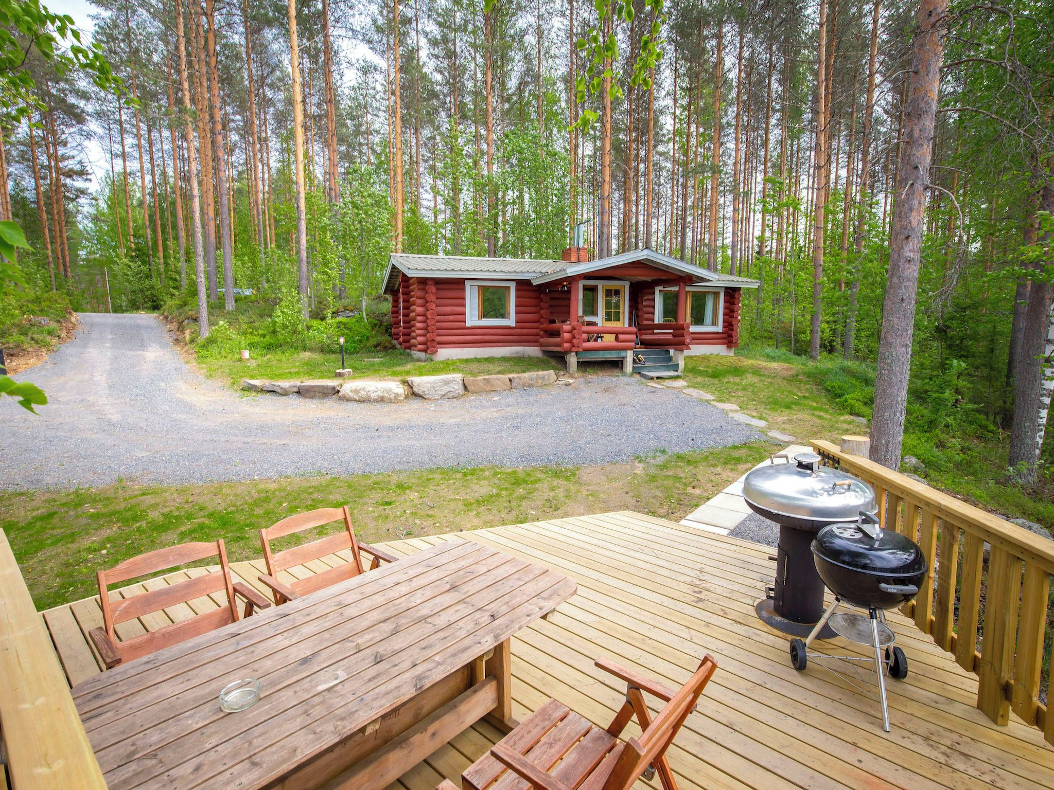 A cabin with a deck, a barbecue grill, and a gravel driveway leading to the entrance.