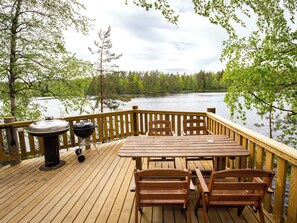 Een houten terras met een tafel en stoelen, uitkijkend op een meer met bomen op de achtergrond.