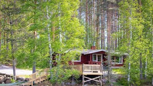 Een chalet met een houten terras en een klein terras, omringd door hoge bomen en een waterpartij.