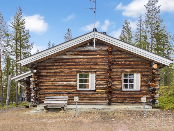 Une cabane rustique en rondins de bois, dotée de deux fenêtres, d’un banc et d’une horloge fixée au mur.