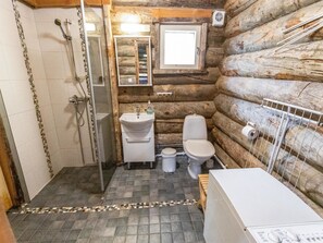 Une salle de bain rustique, aménagée dans une cabane en rondins, avec une douche, un lavabo, des toilettes et une petite fenêtre.