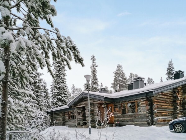 A log cabin with a stone chimney, surrounded by snow-covered trees.