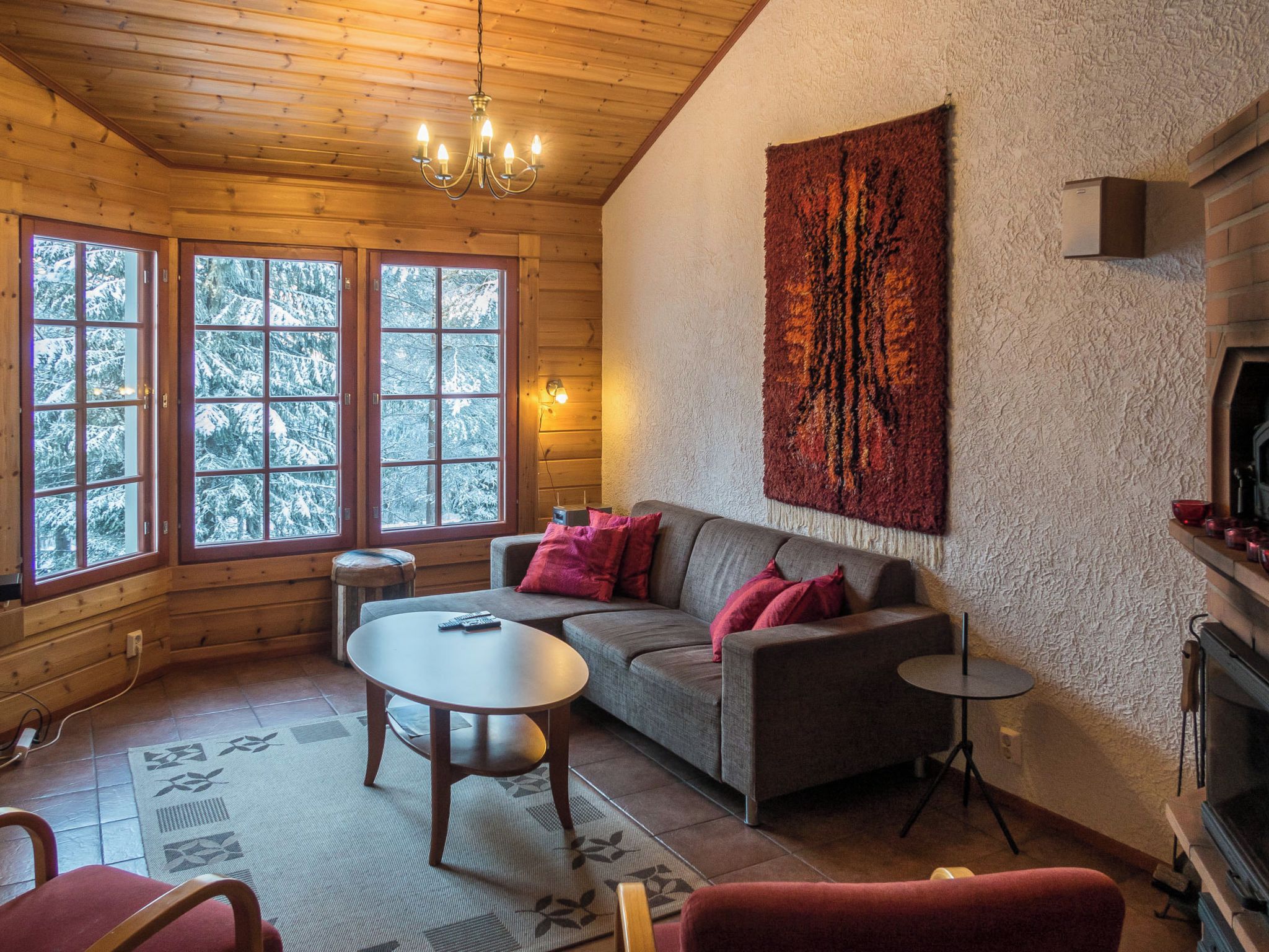 Een gezellig chaletinterieur met houten plafond, een open haard, een bank met kussens, een ronde tafel en een groot raam met uitzicht op besneeuwde bomen.