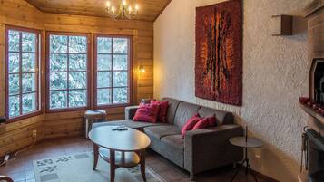 Een gezellig chaletinterieur met houten plafond, een open haard, een bank met kussens, een ronde tafel en een groot raam met uitzicht op besneeuwde bomen.