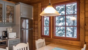 Interior de una cabaña de madera con cocina, mesa de comedor, sillas y un amplio ventanal con vistas nevadas.