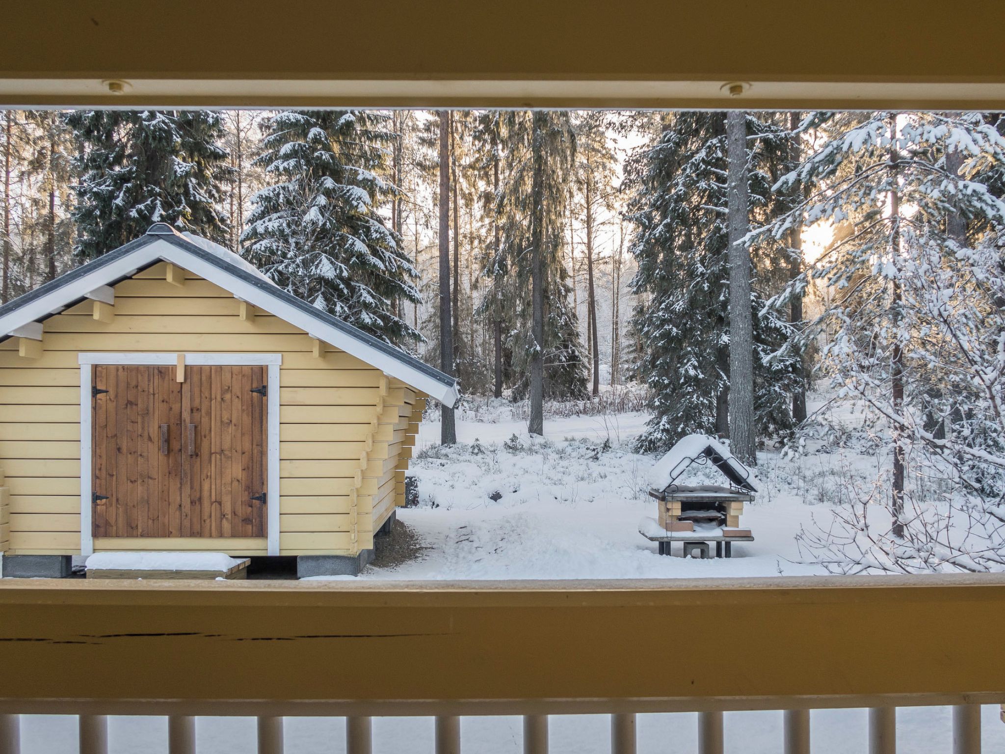 Una cabaña de madera con techo nevado y una estructura pequeña contigua, rodeada por un bosque cubierto de nieve.