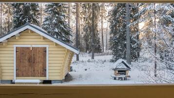 Una cabaña de madera con techo nevado y una estructura pequeña contigua, rodeada por un bosque cubierto de nieve.