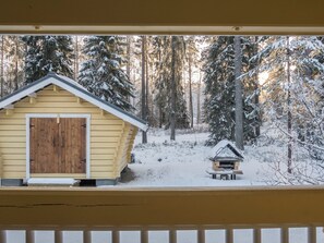 Une cabane en bois, dont le toit est recouvert de neige, et une petite construction adjacente, le tout entouré d’une forêt enneigée.