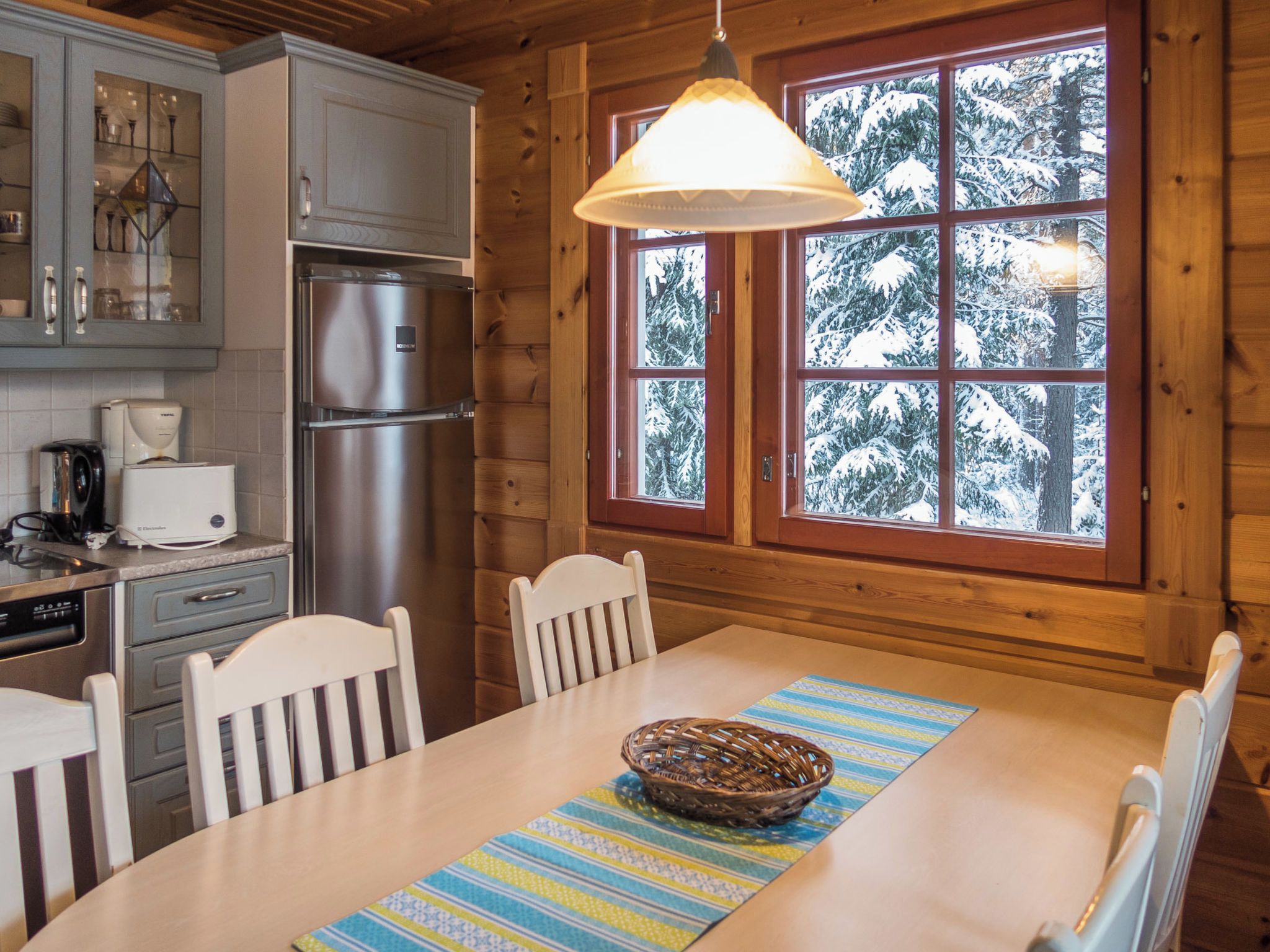 Interior de una cabaña de madera con una mesa de comedor, sillas, un refrigerador, una cafetera y una ventana con vista nevada.