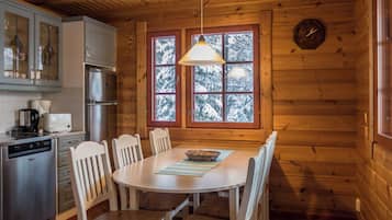 Interior de una cabaña de madera con cocina, una mesa de comedor, sillas y una ventana que ofrece vistas a árboles nevados.