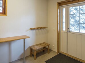 Une pièce avec un banc en bois, une petite table, un porte-manteau et une porte donnant sur des arbres couverts de neige.