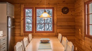 Interior de una cabaña de madera con una mesa de comedor, sillas, una araña y un refrigerador.