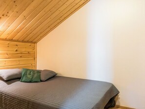 A single bed with a gray bedspread and two pillows, one green and one gray, in a room with wooden ceiling beams and white walls.