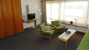 Une chambre d’hôtel avec une armoire en bois, un téléviseur, un bureau avec un ordinateur, deux fauteuils verts, une petite table et une fenêtre avec des rideaux.