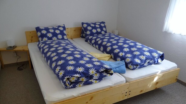 Una cama doble con estructura de madera, dos almohadas estampadas con diseño de margaritas y una colcha azul.