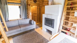 L’intérieur d’une cabane en rondins, avec un canapé gris, une cheminée et une étagère en bois où sont empilés des bûches.