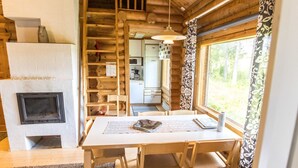 L’intérieur chaleureux d’un chalet avec une table à manger en bois, des chaises, une cheminée, un coin cuisine et une grande fenêtre agrémentée de rideaux.