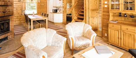 Interior de una cabaña de troncos con comedor, dos sillones, una mesa de centro, chimenea y zona de cocina.