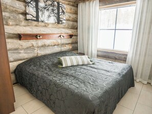 Une chambre avec un mur en rondins de bois, un lit recouvert d’une courtepointe à motifs, une étagère en bois et une fenêtre avec des rideaux.