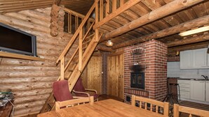 A log cabin interior with a wooden staircase, a brick fireplace, a dining table, and a kitchen area.