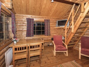 L’intérieur d’un chalet en bois comprend une table à manger, deux chaises, un téléviseur et une fenêtre avec des rideaux.