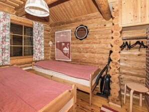 Une cabane en bois avec deux lits, une fenêtre ornée de rideaux à motifs floraux, une lampe fixée au mur et un aspirateur.
