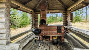 Een houten terras met een bakstenen open haard en een barbecue, omgeven door een houten omheining en bomen.