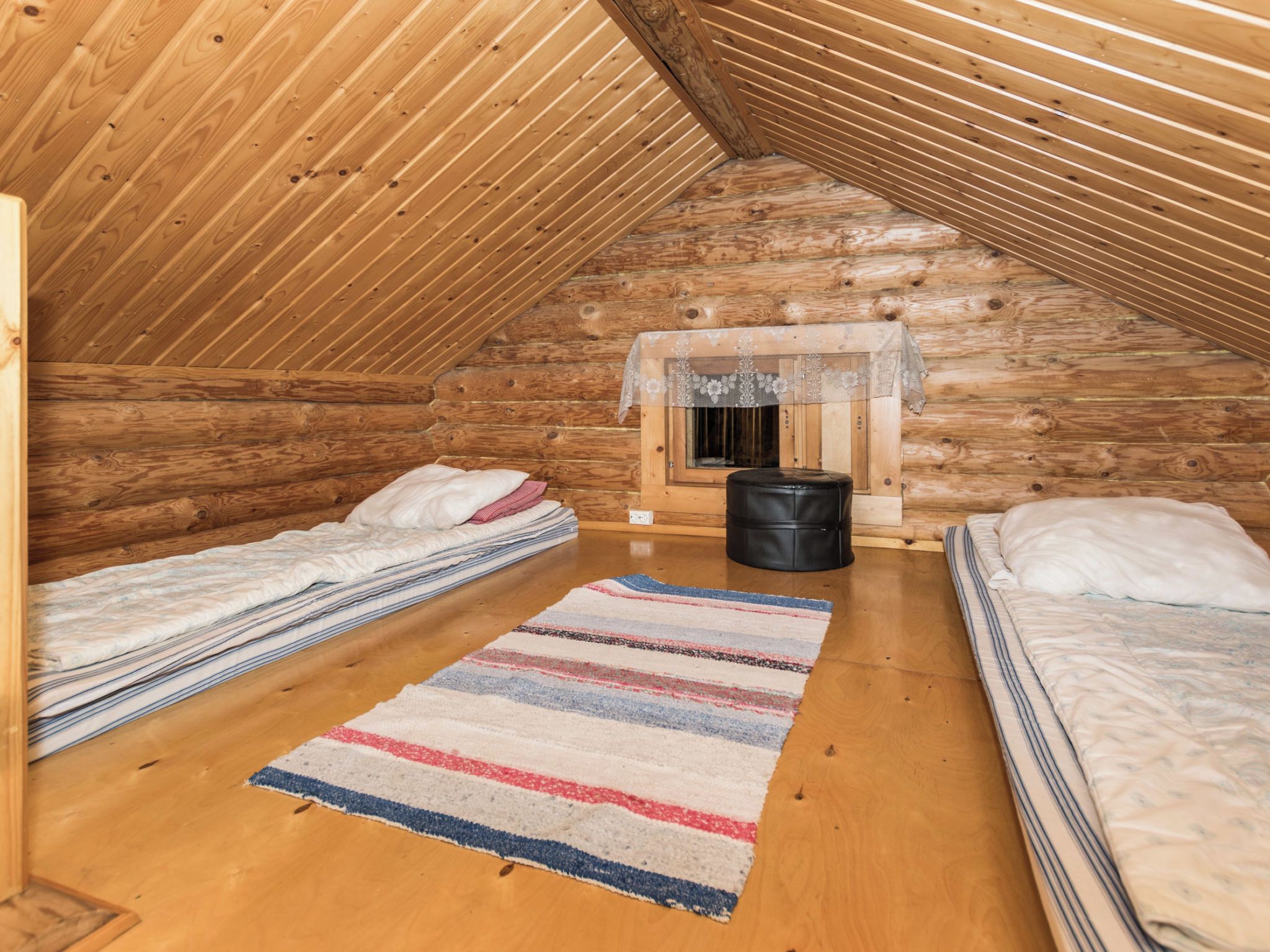 Tunnelmallinen ullakkohuone, jossa on puuseinät, kaksi sänkyä, ikkuna ja musta roskakori.