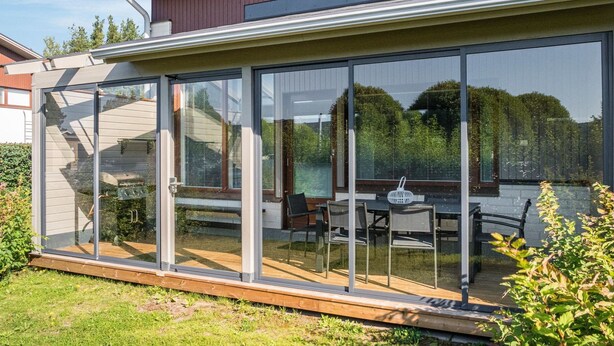 Une maison moderne dotée d’un grand patio vitré, d’une terrasse en bois et de mobilier d’extérieur.