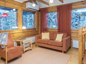 Interior acolhedor de cabana com sofá, poltrona, luminária e janela com vista para árvores cobertas de neve.