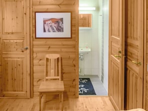 A wooden room with a chair, a framed picture on the wall, and a view into a bathroom with a sink and a tiled wall.