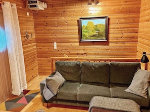 A wooden cabin interior with a sofa, a framed picture, and a window with a view of trees.