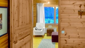 A wooden cabin room with a bed, a white chair, a small table with a glass object, and a window with curtains.