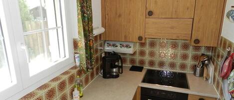 Een kleine keuken met houten kastjes, een inbouwoven, een koffiezetapparaat en een raam met gordijnen.